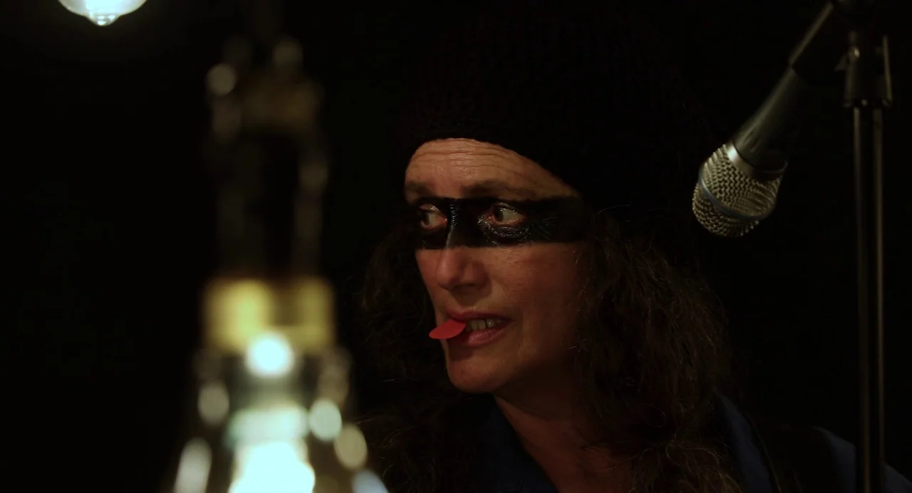 Hellbender (2021) movie still: closeup — A dark space features a person wearing a black mask around their eyes. The subject holds a red guitar pick…