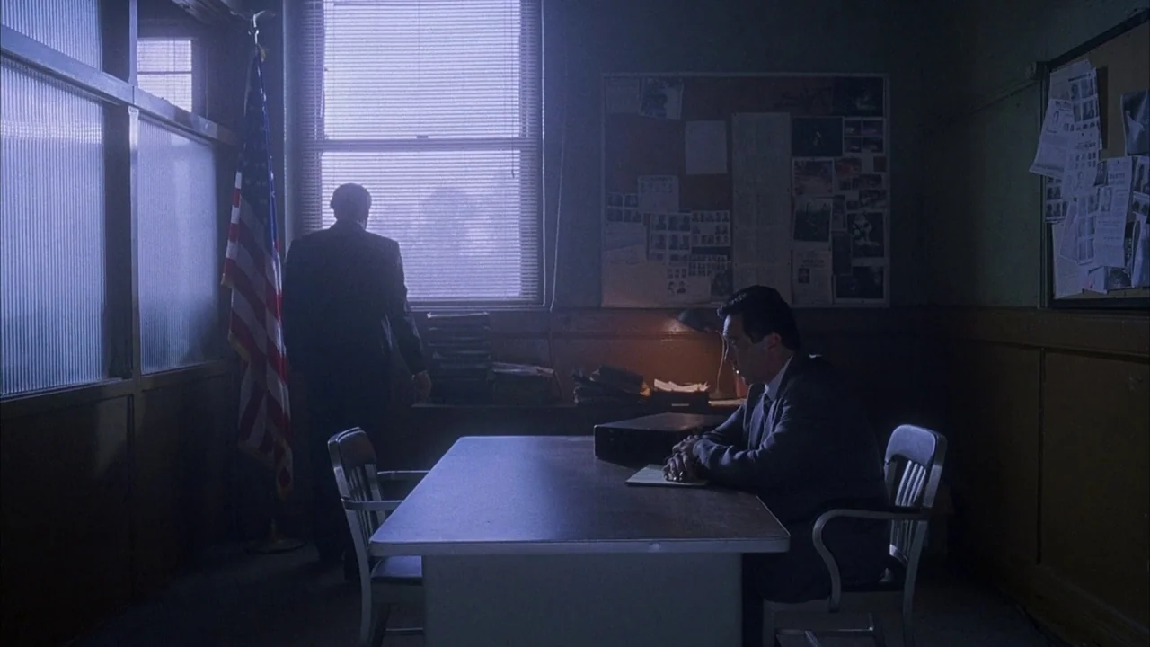 Saw movie still: wide — A dark, spartan room with a corkboard covered in papers and a metal table. One man stands looking out the window while the…