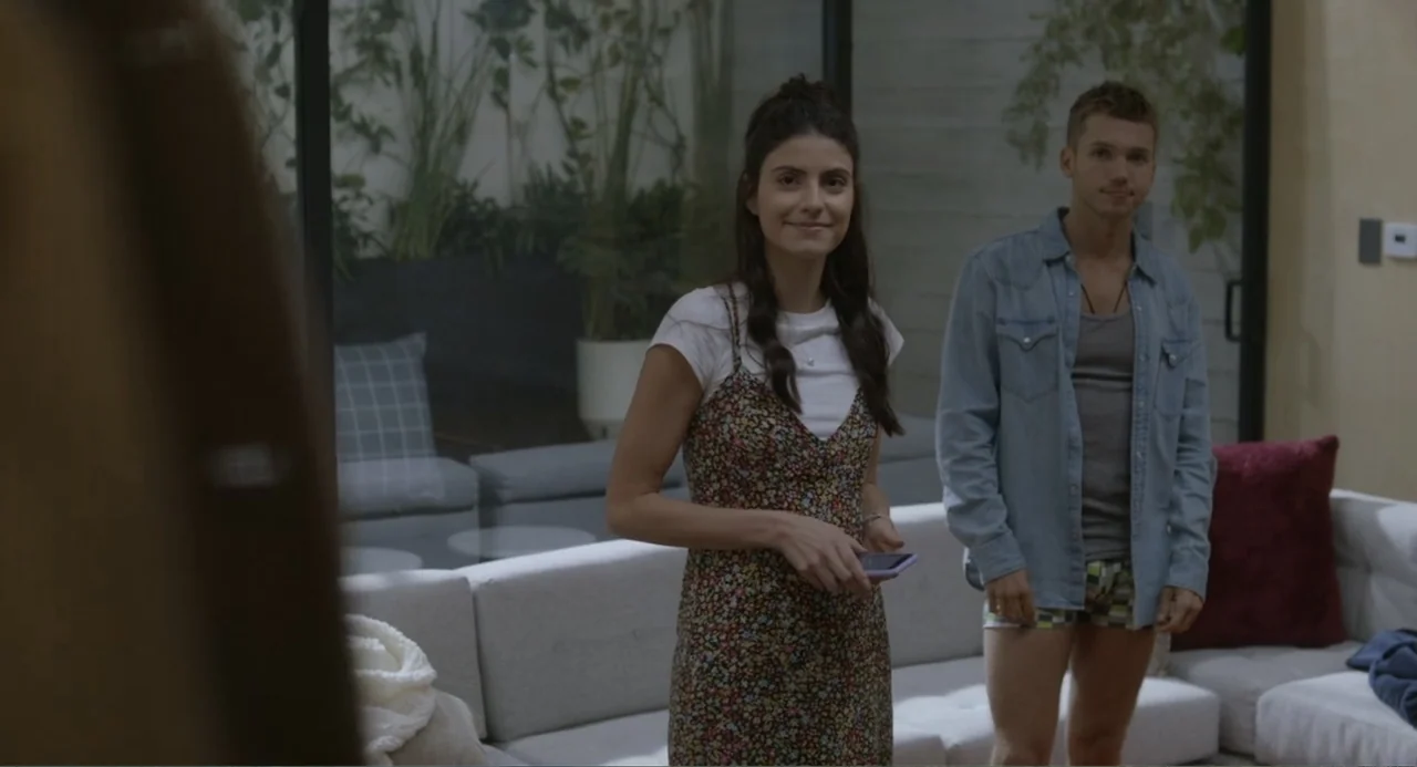 Straight (2024) movie still: medium — A modern living room with a large glass wall featuring an interior courtyard plant display. A young woman and…