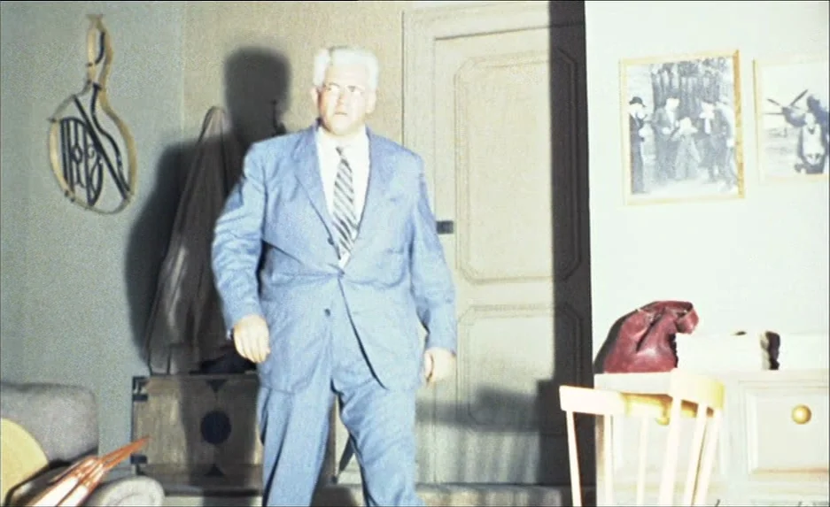Rear Window (1954) movie still: fullbody — A furnished interior features beige walls adorned with framed pictures and a door. A stout man in a blue…