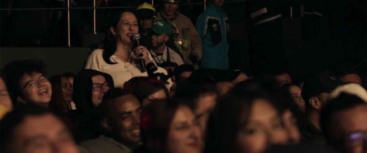 Making of: F*CKS NEWS (2025) movie still: medium — A dimly lit auditorium is filled with a seated audience in darkness. One woman stands in the…