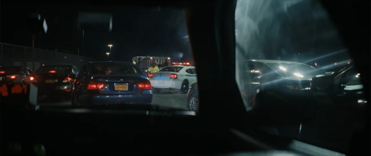 Daddio (2024) movie still: wide — A nighttime road scene shows a line of cars stopped behind an emergency vehicle. Bright red brake lights reflect…