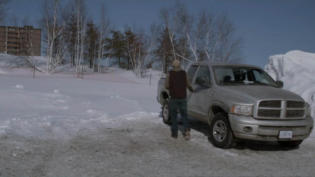 The Captive (2014) movie still: fullbody — A snowy outdoor lot features a silver pickup truck parked near leafless birch trees. A man in a dark vest…