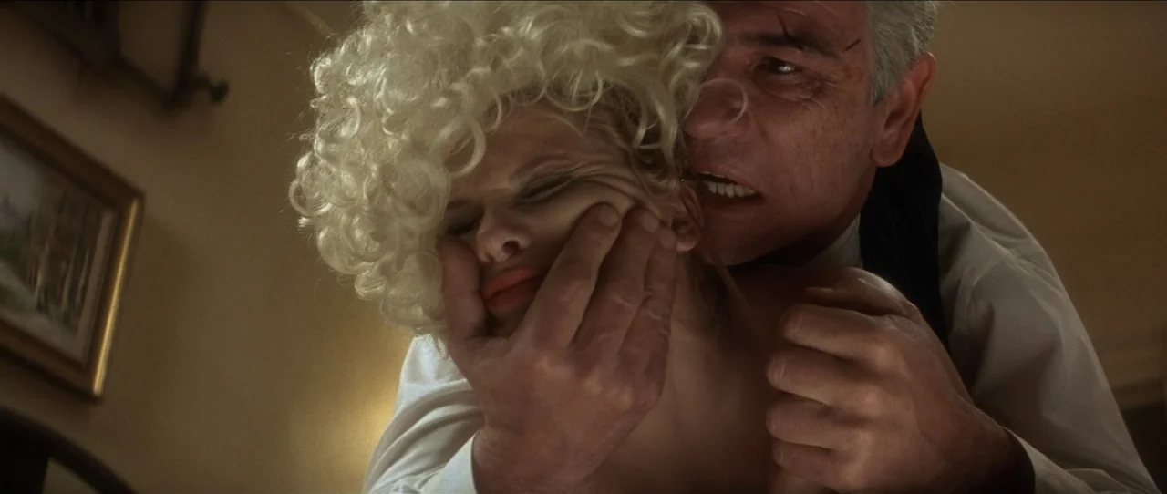 Cobb (1994) movie still: closeup — A dimly lit room with neutral wall tones and a framed picture visible. A man clutches the face of a blonde woman…