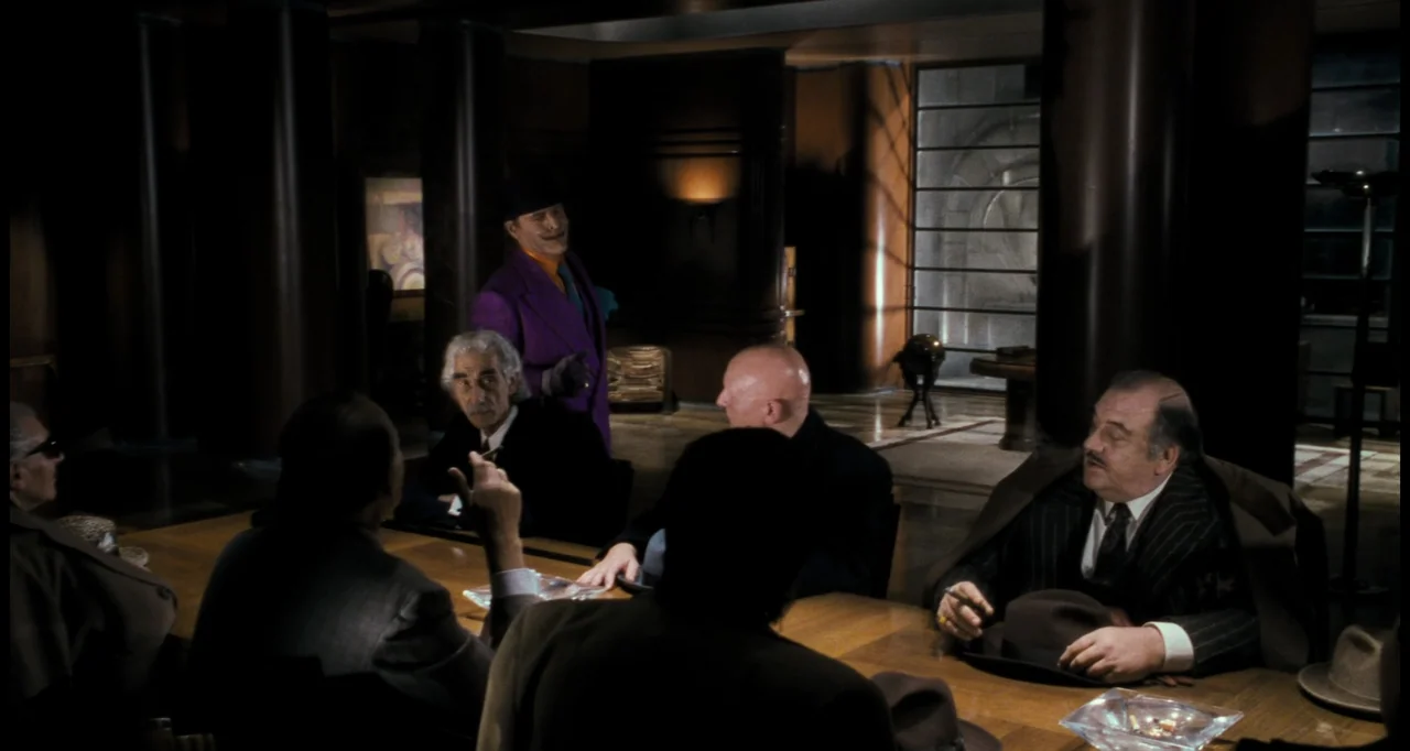 Batman (1989) movie still: medium — A dimly lit corporate boardroom features dark wood paneling and a long conference table. A man in a purple suit…