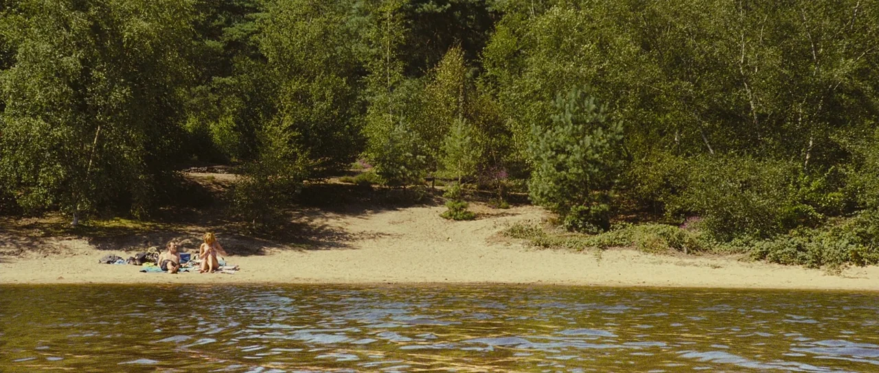 Eden Lake (2008) movie still: wide — A secluded sandy bank is bordered by dense green foliage and calm water. A man and a woman lie together on the…