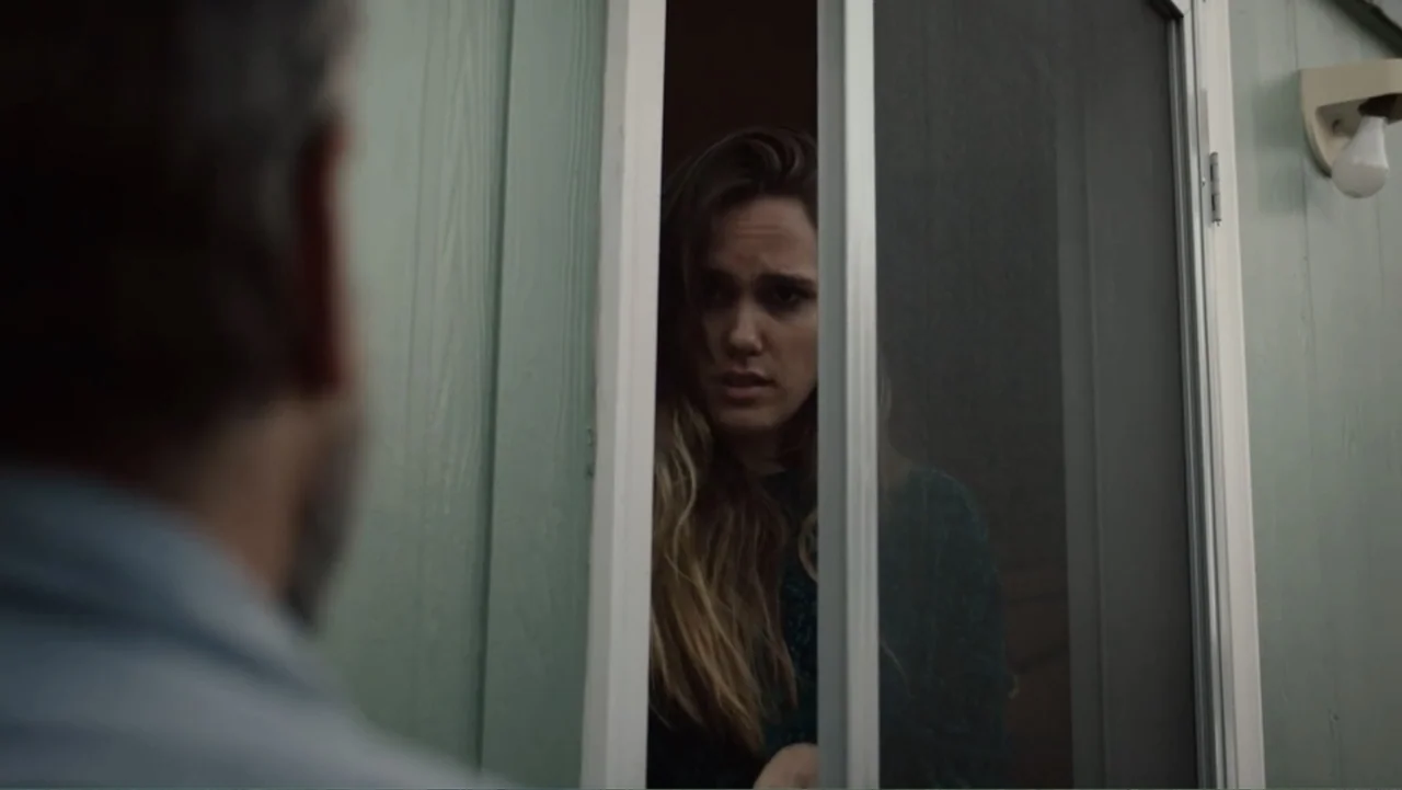 The Cleaner (2021) movie still: medium — A woman stands behind a screen door of a light green sided house. A man stands in the foreground, partially…