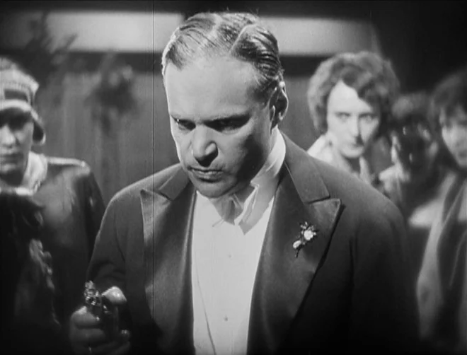 Pandora’s Box (1929) movie still: medium — A crowded social gathering occurs within a dimly lit, large indoor space. A man in formal attire stands…