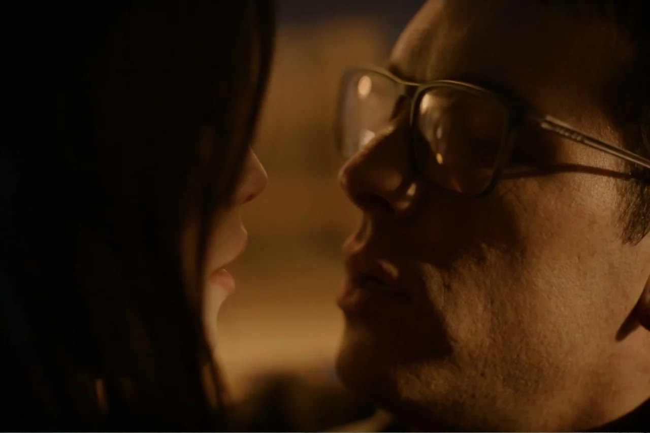 Cross the Line (2020) movie still: closeup — A darkened interior space features two subjects facing each other closely. The man wears glasses while…