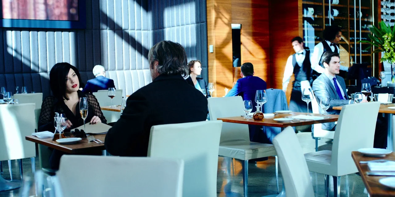 Polar (2019) movie still: medium — A modern restaurant with wooden paneled walls and floor-to-ceiling padded blue acoustic wall panels. A man and…