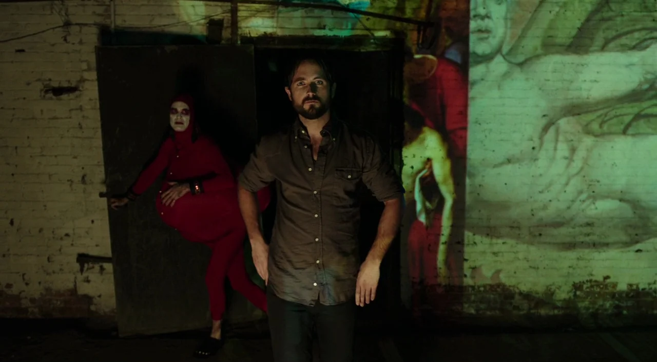 Urge (2016) movie still: medium — A dark brick wall basement serves as the backdrop for a projection display. A man stands in front of the projector…