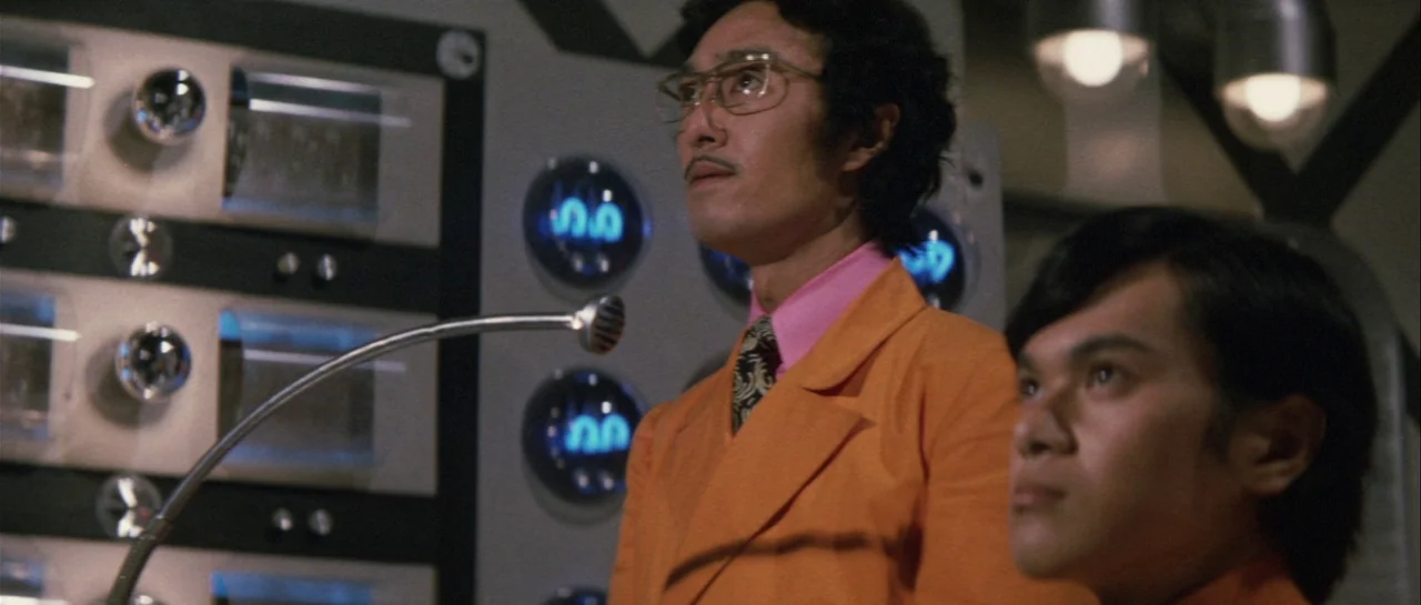 Godzilla Vs Gigan (1972) movie still: medium — A science facility interior features complex equipment with glowing blue nixie tube displays. Two men…