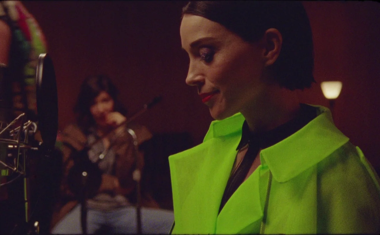 The Nowhere Inn (2020) movie still: closeup — A dimly lit room contains recording equipment with warm ambient light in the background. A woman in a…