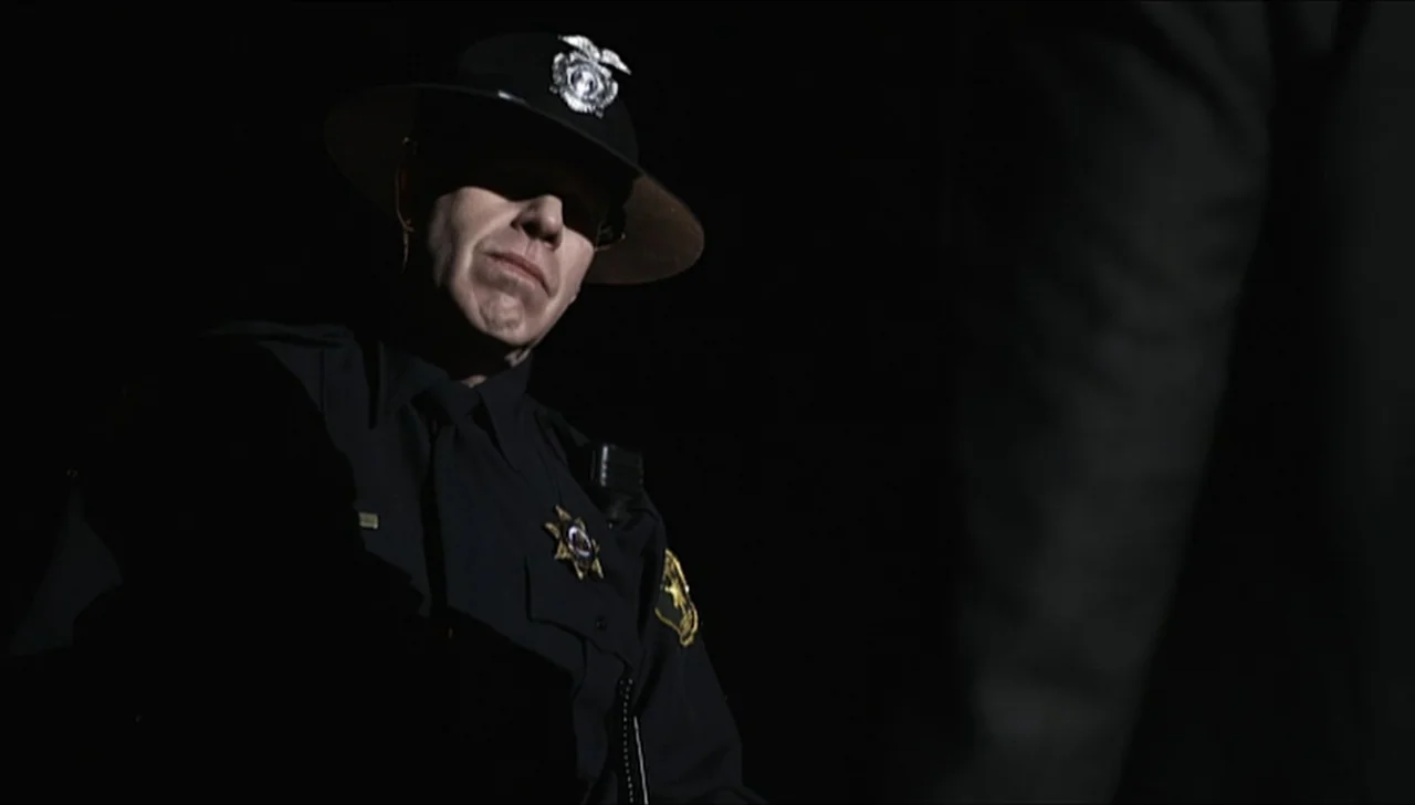 Dark Country (2009) movie still: medium — A dark night setting featuring a police officer partially obscured by shadow. The officer stands facing an…