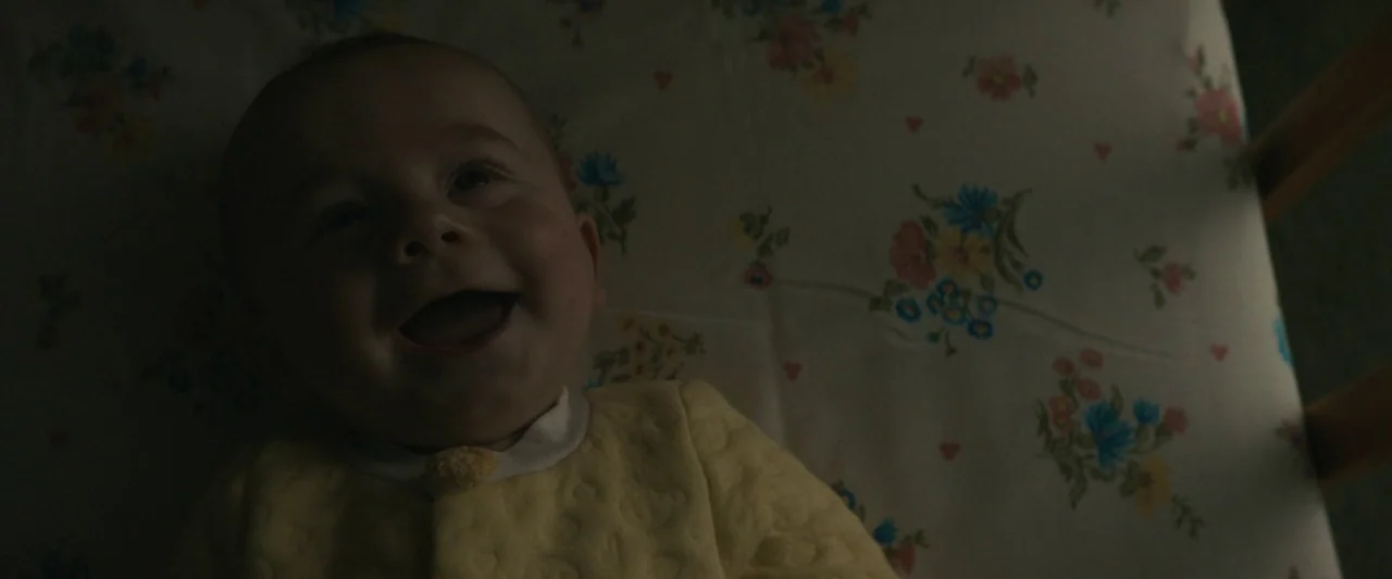 I'm Your Woman (2020) movie still: closeup — A baby rests on a sheet with a vintage floral pattern in a crib. The infant lies on its back and smiles…