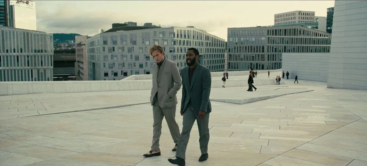Tenet (2020) movie still: fullbody — A sprawling stone rooftop plaza with geometric tiled surfaces and modern buildings in distance. Two men walk…