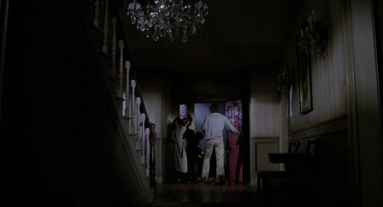 The Amityville Horror (1979) movie still: wide — A dimly lit residential foyer features a wooden staircase on the left side. Several individuals are…