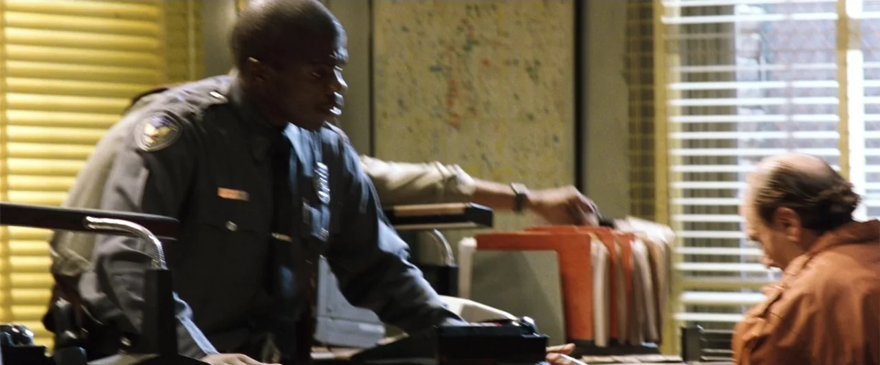 Fallen (2004) movie still: medium — An office interior features desk clutter, hanging file folders, and window blinds. A police officer in uniform…