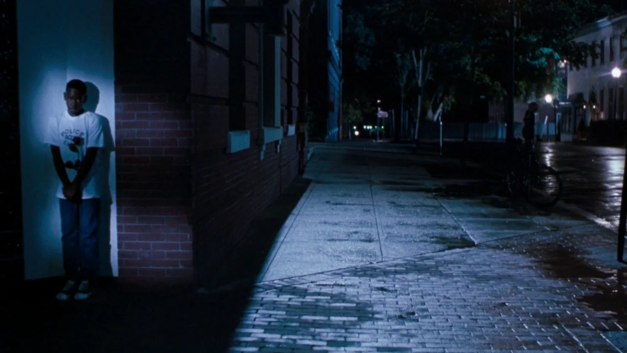 The Exorcist 3 (1990) movie still: fullbody — A nighttime city sidewalk corner features a red brick wall and dark pavement. A young man stands in a…