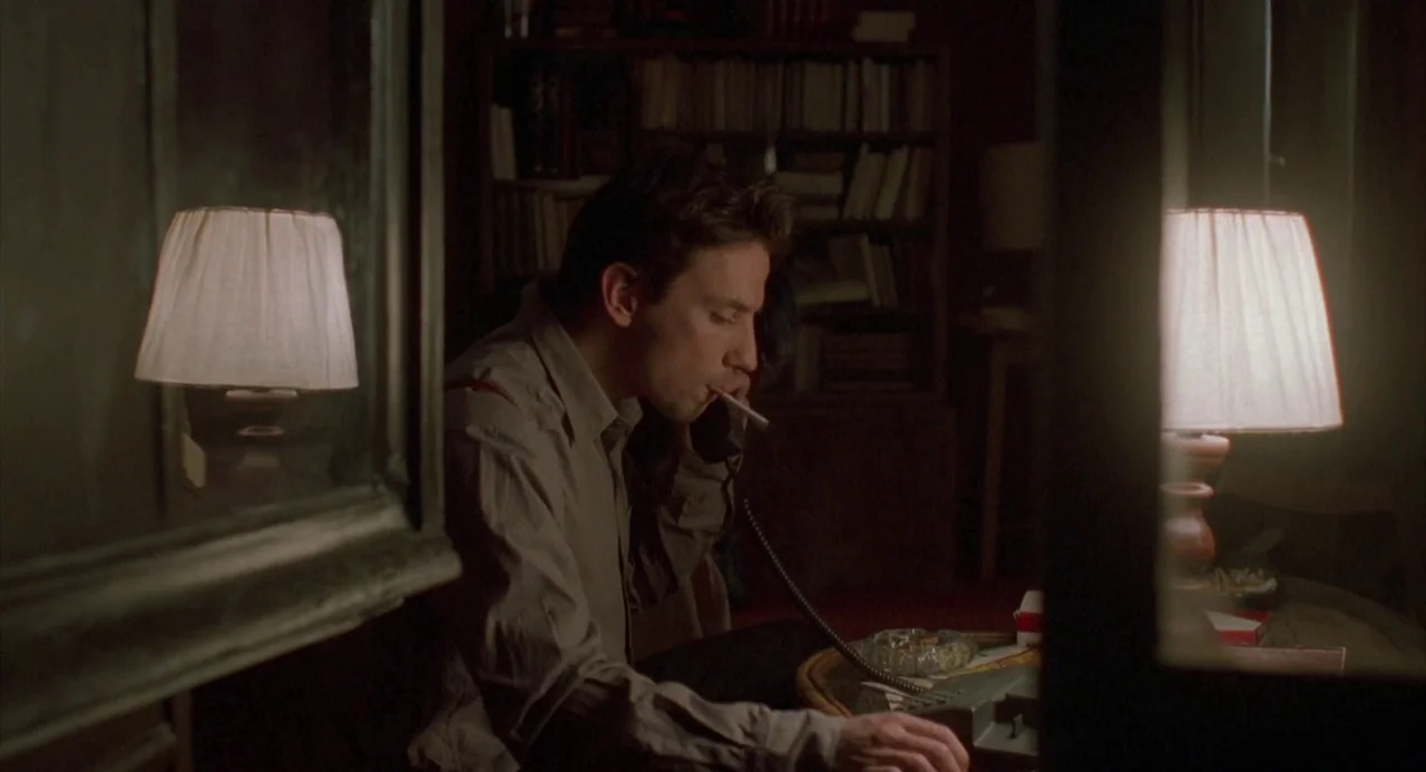 Three Colours: Red (1994) movie still: medium — A dimly lit room contains a bookshelf and a small table with a lamp. A man sits at the table,…