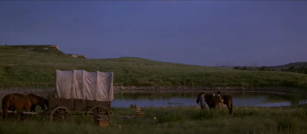 Ride With The Devil movie still: wide — A grassy landscape features a reflective pond under a fading deep blue evening sky. A covered wagon pulled…