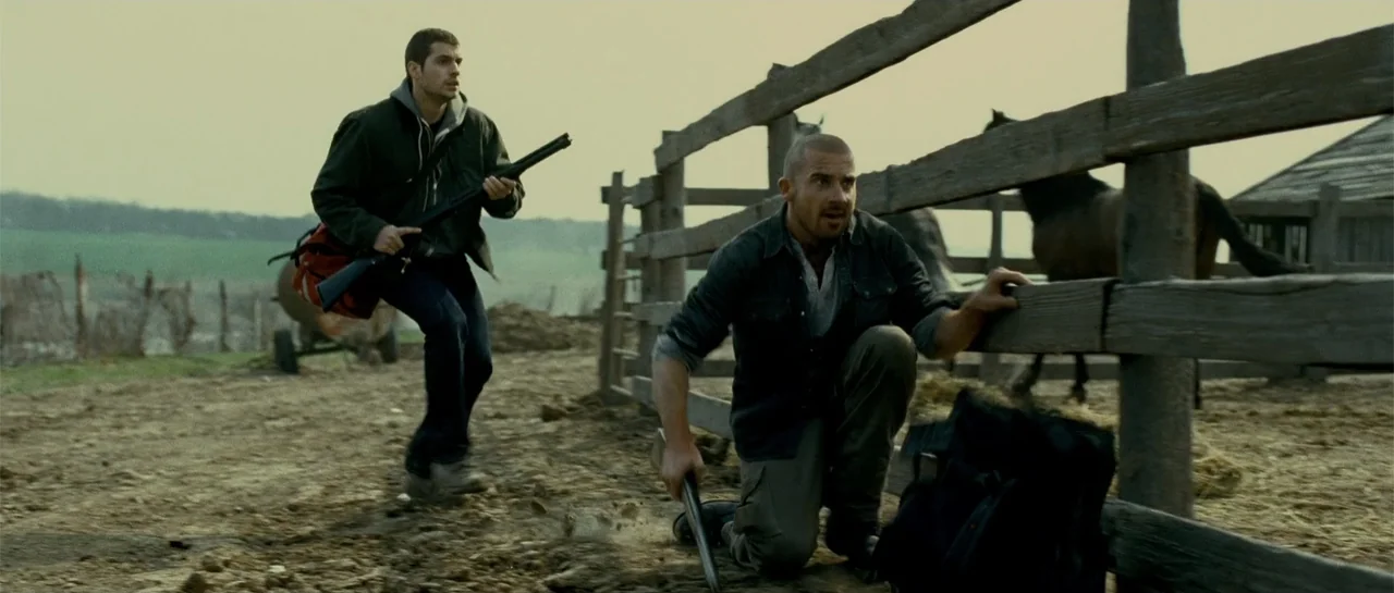 Blood Creek (2009) movie still: fullbody — A rural dirt enclosure features a weathered wooden fence and horses in the background. Two armed men…