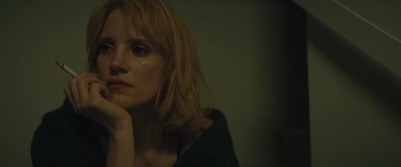 A Most Violent Year (2014) movie still: closeup — A woman sits on a dimly lit indoor staircase with dark walls. She holds a lit cigarette between…