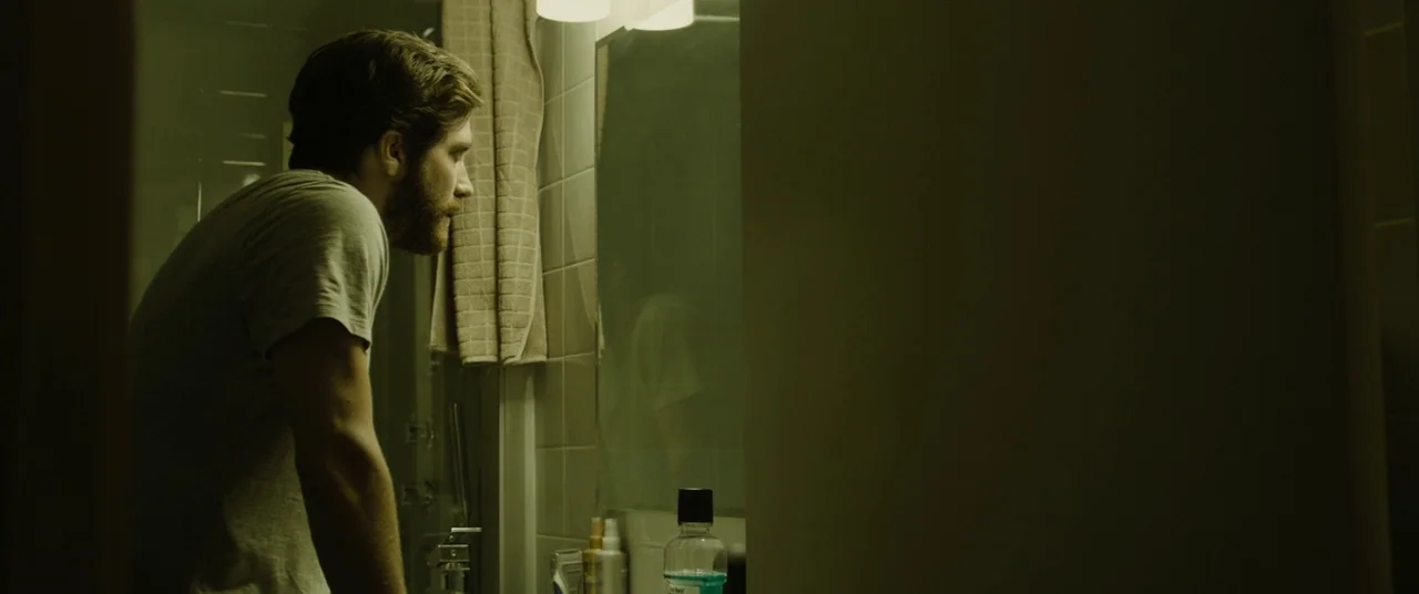 Enemy (2013) movie still: medium — A small residential bathroom featuring beige wall tiles and a vanity mirror. A man stares at his reflection while…