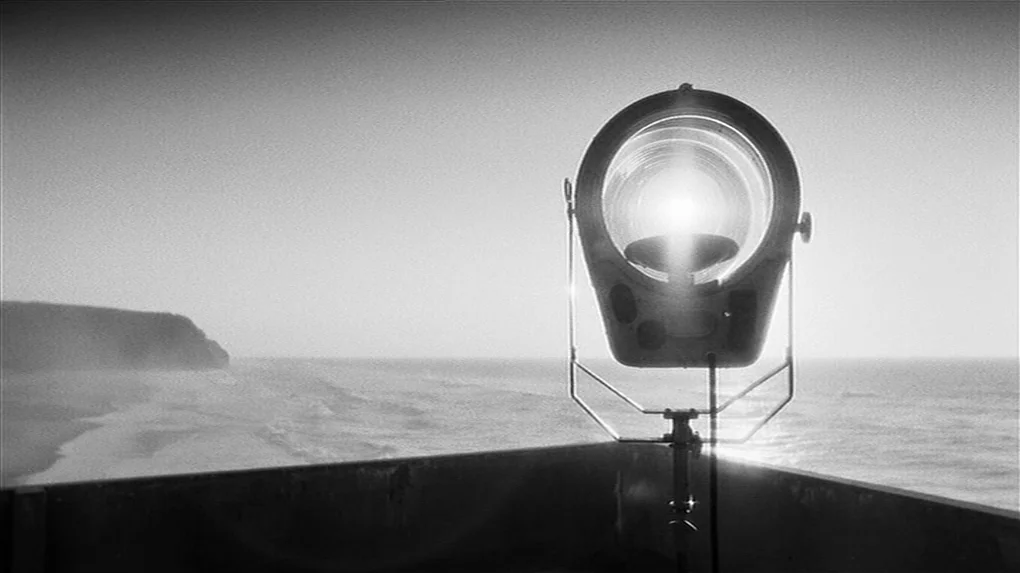 The State of Things (1982) movie still: wide — A metal spotlight stands on a concrete balcony overlooking a calm ocean beach. The sky is clear and…