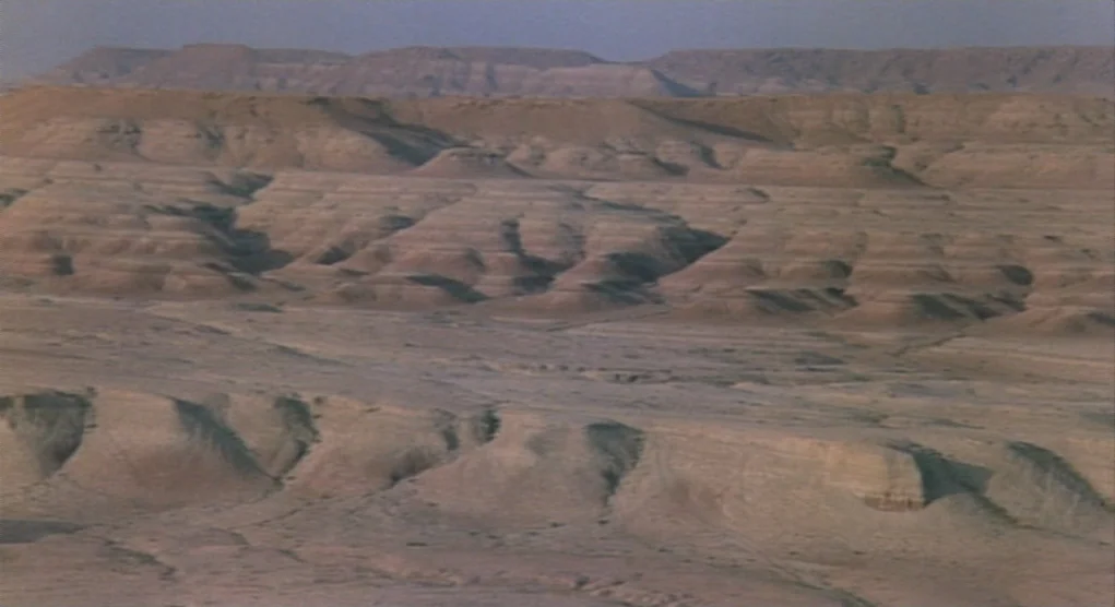 Time Bandits (1981) movie still: establishing — An arid, eroded desert landscape stretches toward a series of distant, weathered plateaus. The…