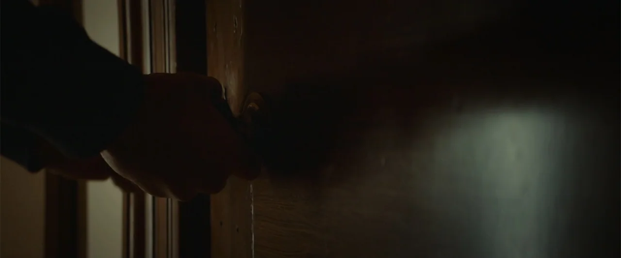 The Cellar (2022) movie still: closeup — A close-up view shows a hand gripping a metallic doorknob on a wooden door. The surrounding area remains in…