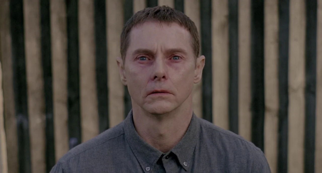 Possum (2018) movie still: closeup — A man stands in front of a rustic fence made of vertical wooden slats. The subject looks directly toward the…