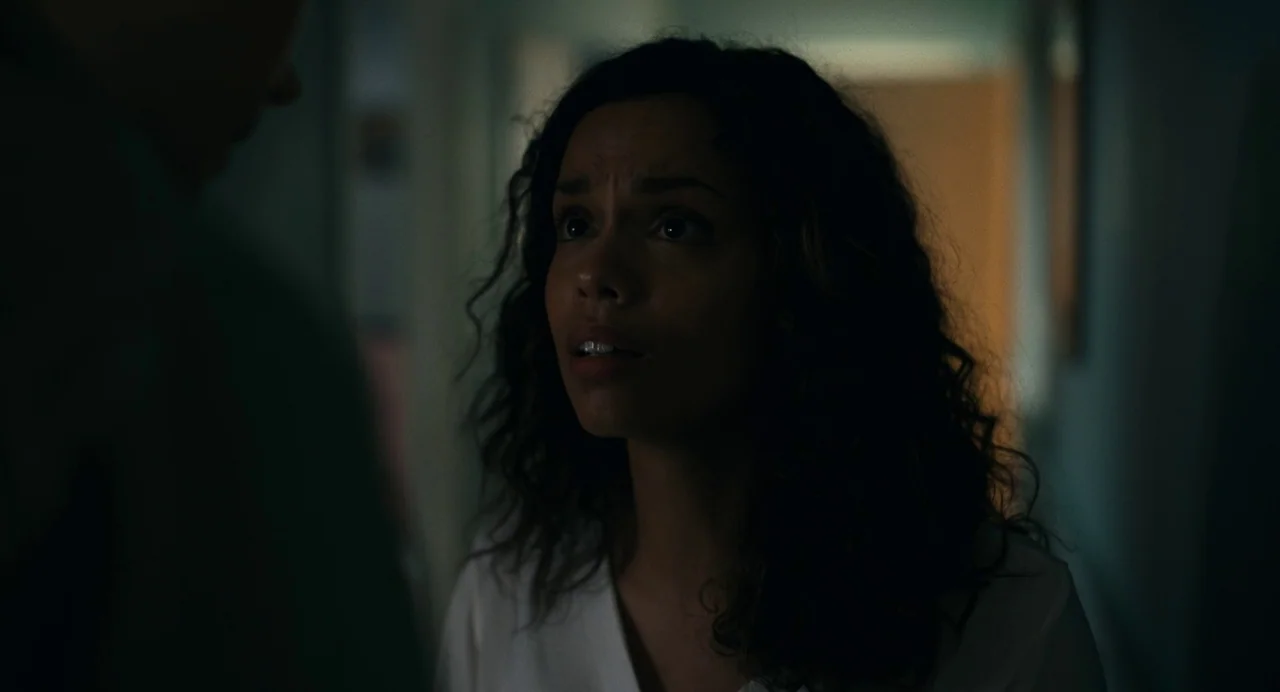 Barbarian (2022) movie still: closeup — A dimly lit indoor hallway with a blurred figure in the foreground. A woman with curly hair stands in the…