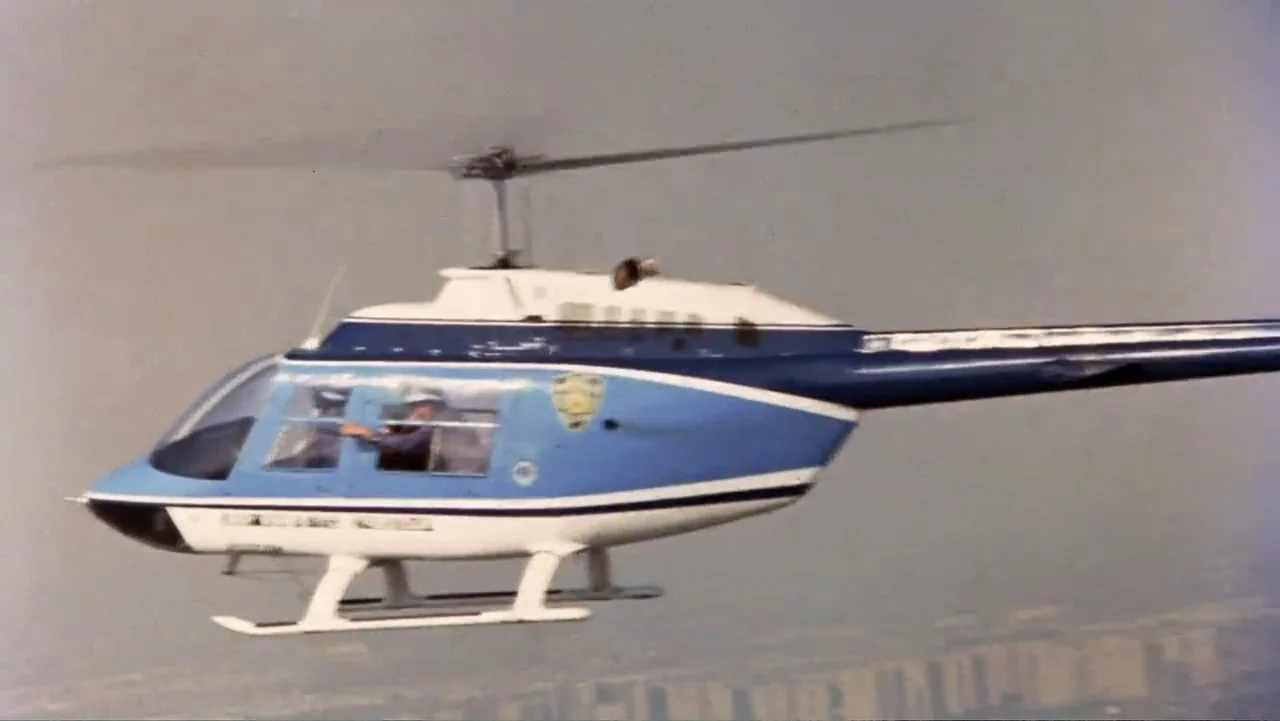Dog Day Afternoon (1975) movie still: wide — A blue and white police helicopter flies through a pale, hazy sky. The pilot occupies the cockpit of…