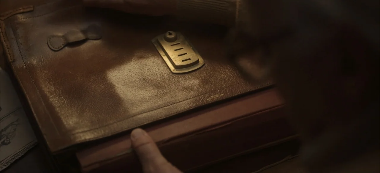One Life (2023) movie still: closeup — A brown leather briefcase with a gold combination lock rests on a dark surface. A person's hand reaches out…
