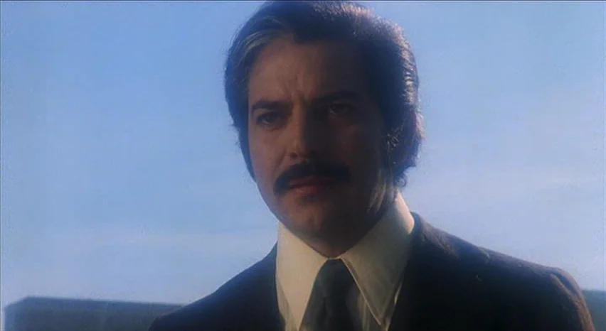 Lisa and The Devil (1973) movie still: closeup — A man stands outdoors against a clear blue sky. He wears a formal suit and looks toward an unseen…