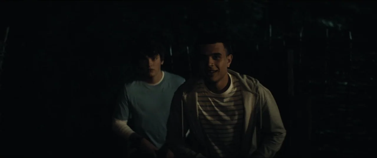 Bonus Track (2024) movie still: medium — An outdoor area at night with minimal illumination from a source in front. Two teenage boys stand close…