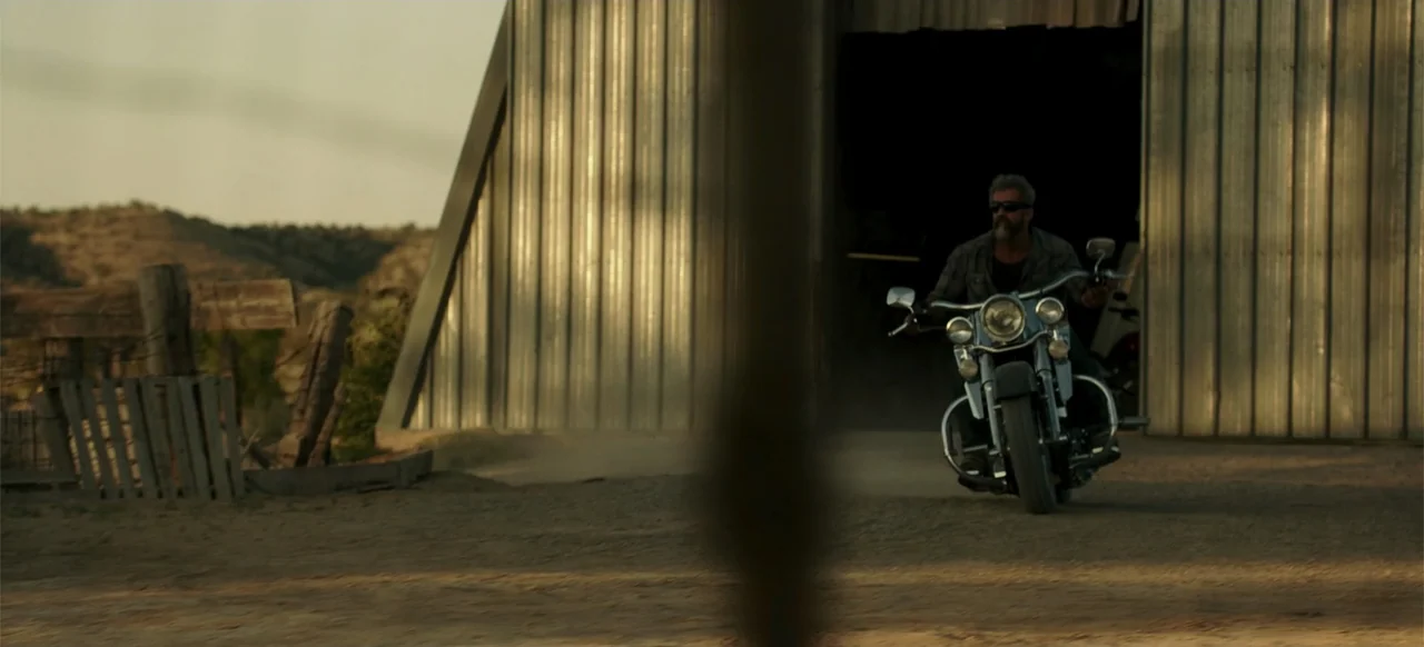 Blood Father (2016) movie still: wide — A man rides a motorcycle out of a large metal barn in a dry field. The man wears sunglasses and a casual…