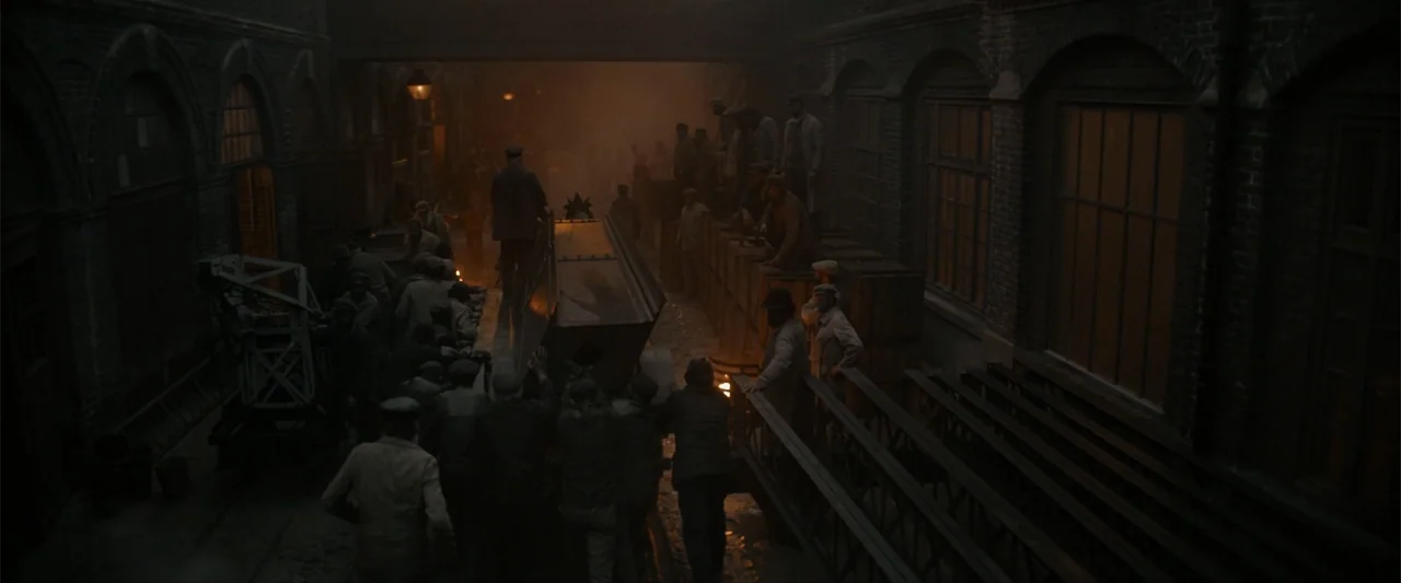 Eiffel (2021) movie still: wide — An industrial brick loading bay at night filled with heavy smoke and dark shadows. A large group of men in period…