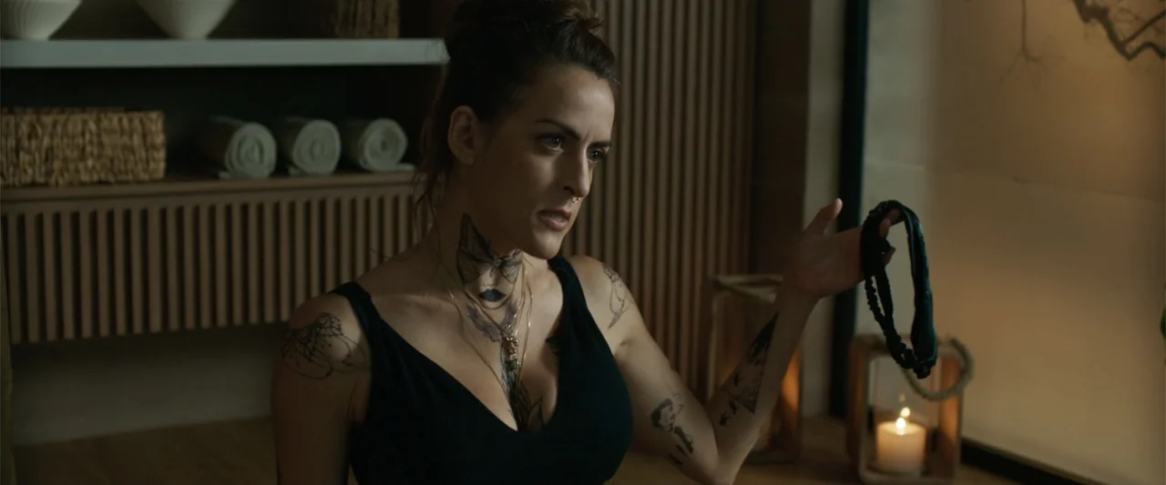 Férias Trocadas (2024) movie still: medium — A dimly lit room with textured wooden walls and shelves holding rolled towels. A woman with visible…