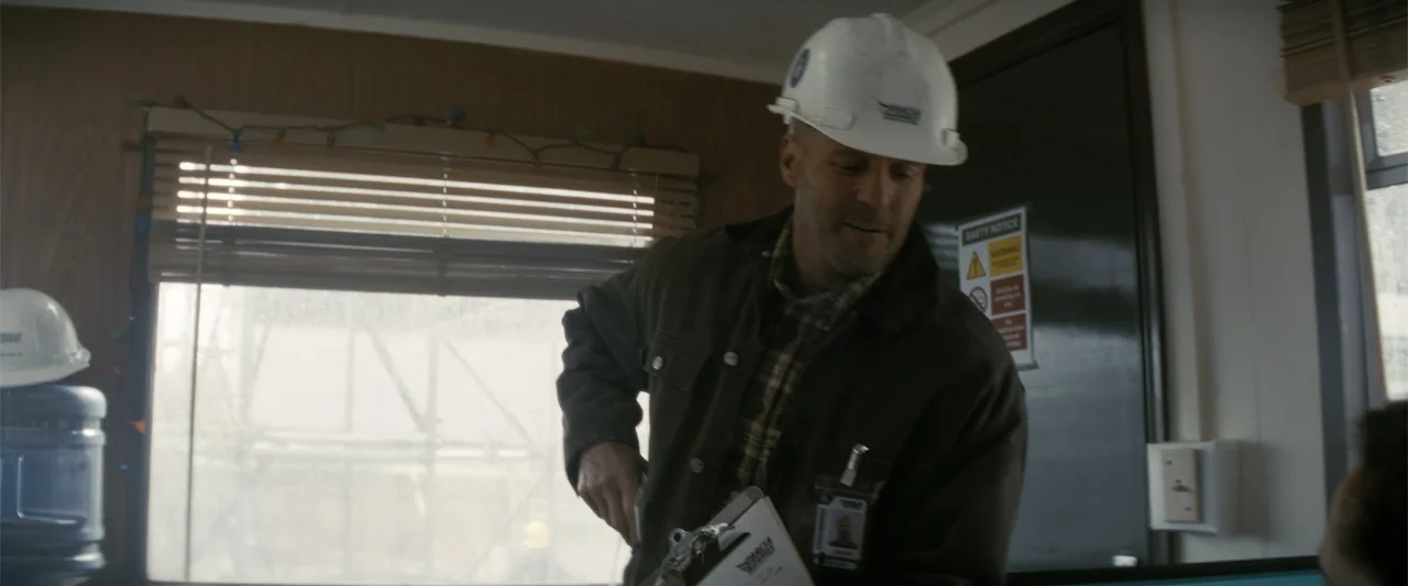 A Working Man (2025) movie still: medium — A small construction trailer interior with wood-paneled walls and a window with blinds. A man in a work…