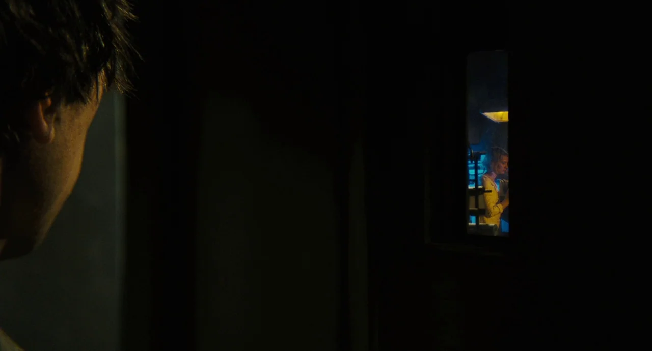 The Double (2013) movie still: medium — A dark, unlit corridor frames a view into an adjacent brightly lit room. A woman stands in the distance…