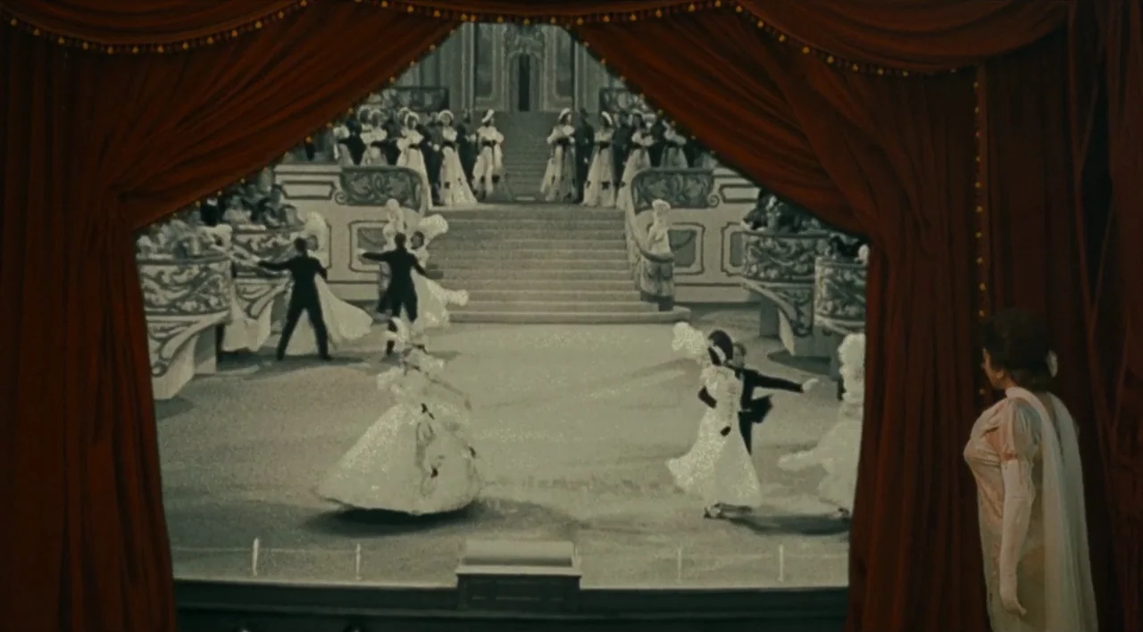 The Other Conquest (1999) movie still: wide — A woman stands in the wings of a theater looking at a staged ball. Dancers in formal attire waltz on…