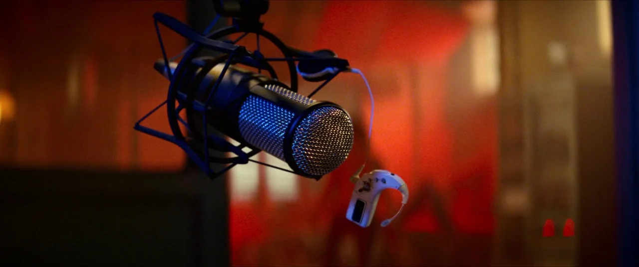 A Quiet Place Part II (2020) movie still: closeup — A close-up of a studio microphone suspended in a dimly lit recording booth. A small hearing aid…