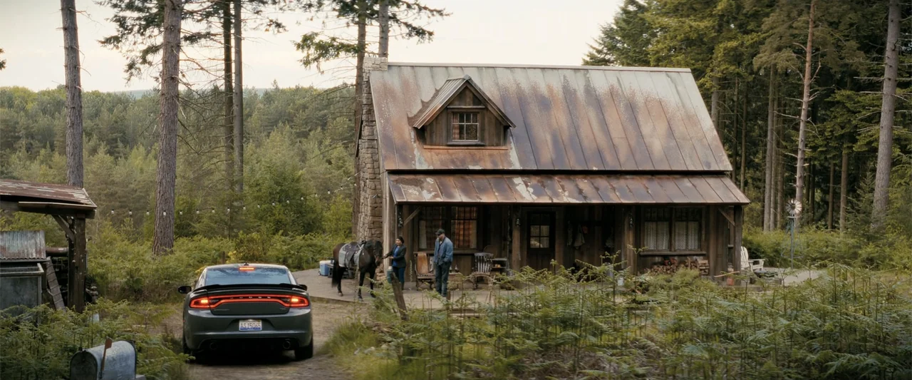 A Working Man (2025) movie still: wide — A weathered rustic cabin sits nestled among pine trees with a parked sedan nearby. A man and a woman stand…