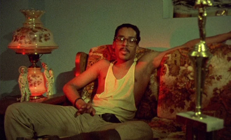 Paris Is Burning (1990) movie still: medium — A 1980s living room features a patterned sofa next to an illuminated floral lamp. A man with glasses…