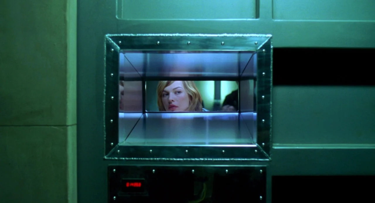 Resident Evil (2002) movie still: closeup — A woman looks through a rectangular reinforced metal viewport on a massive green door. The surrounding…