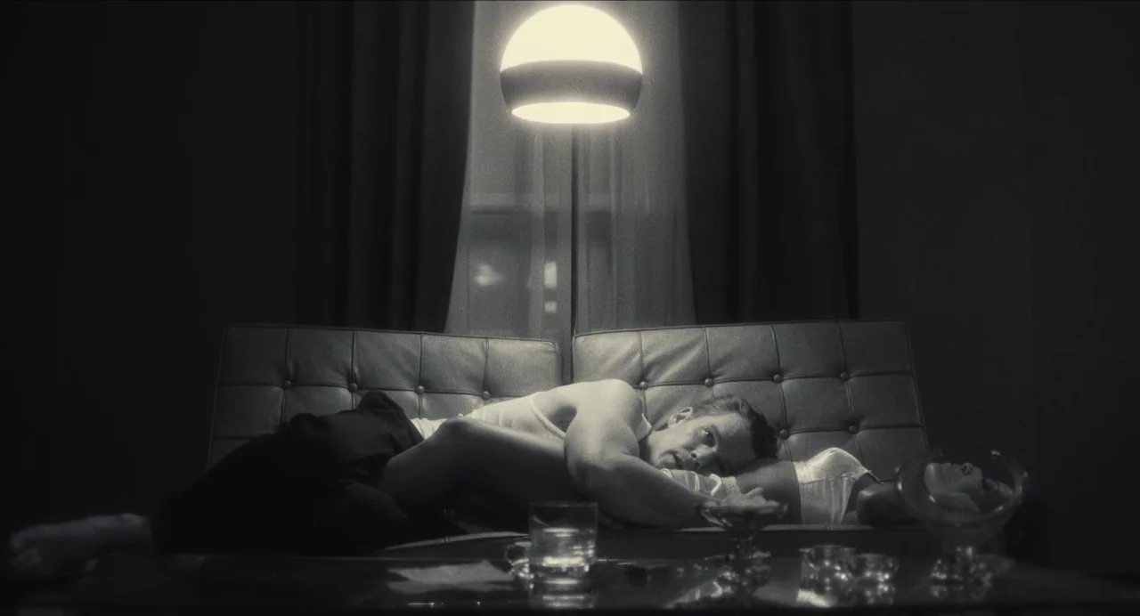 Born To Be Blue (2015) movie still: medium — A dim room features a tufted leather sofa positioned before a window with curtains. A man and a woman…