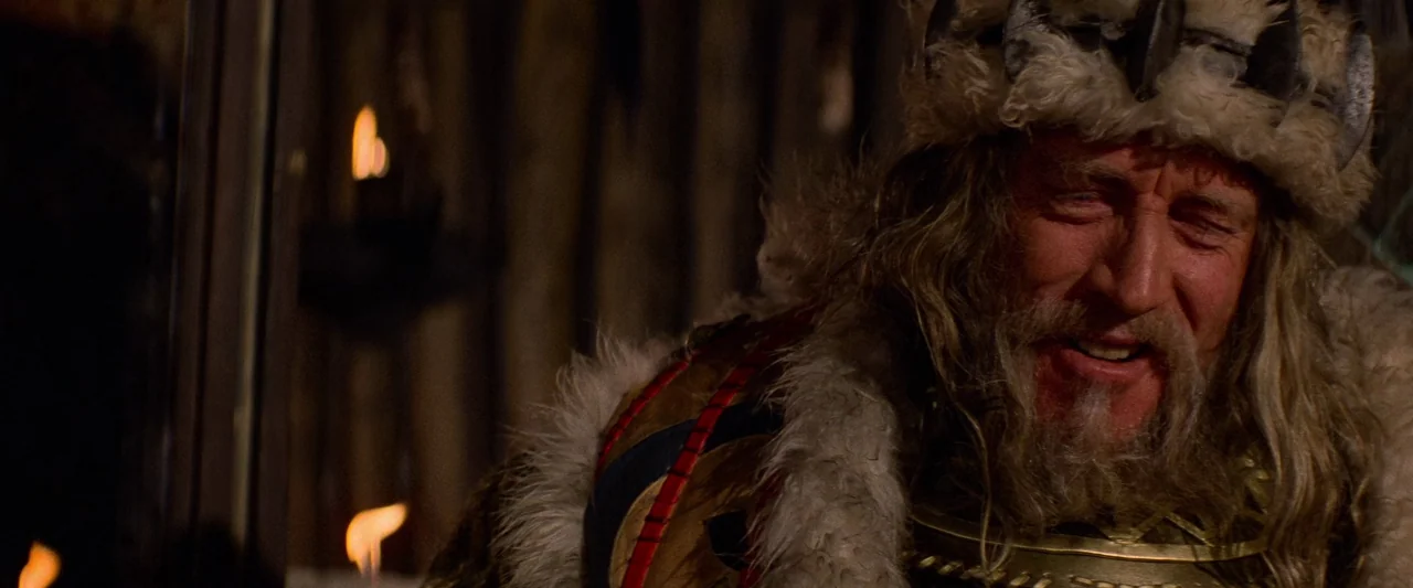 Conan The Barbarian (1982) movie still: closeup — A dimly lit room with wooden walls and flickering flames reflected in the shadows. An older man…
