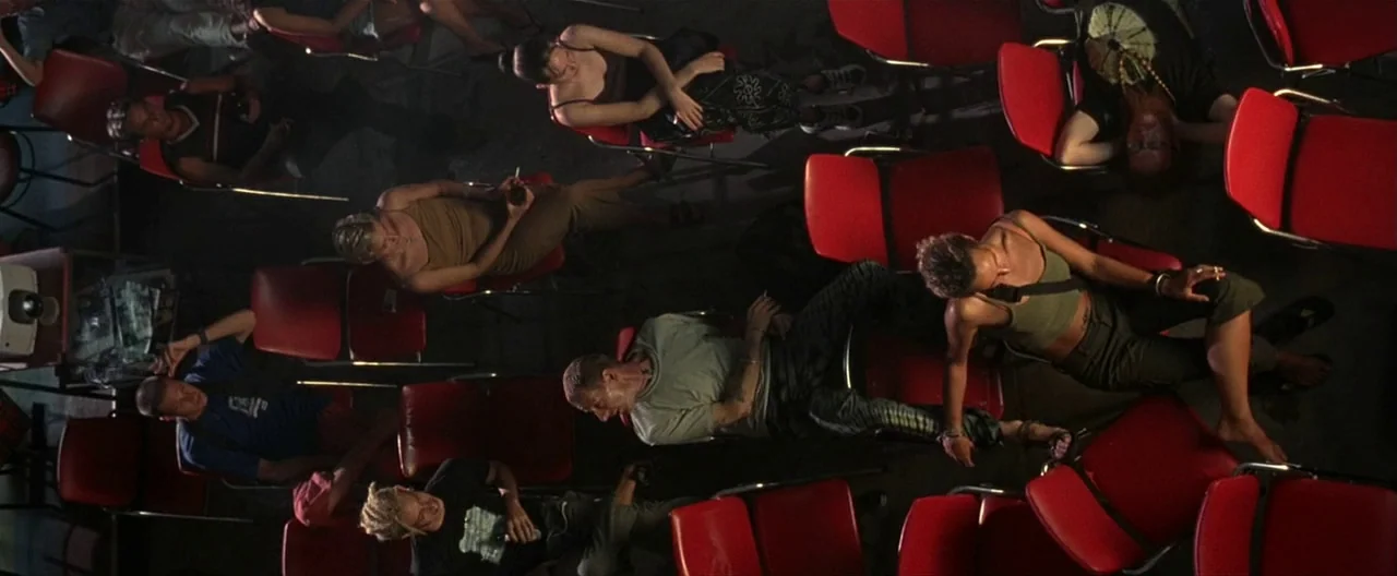 The Beach (2000) movie still: wide — A room with rows of empty and occupied red plastic chairs on a floor. A group of people are scattered across…